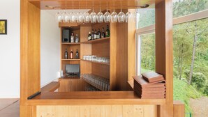 Uma área de bar com balcão de madeira, prateleiras cheias de garrafas e copos, um computador e vista para árvores através de uma janela.