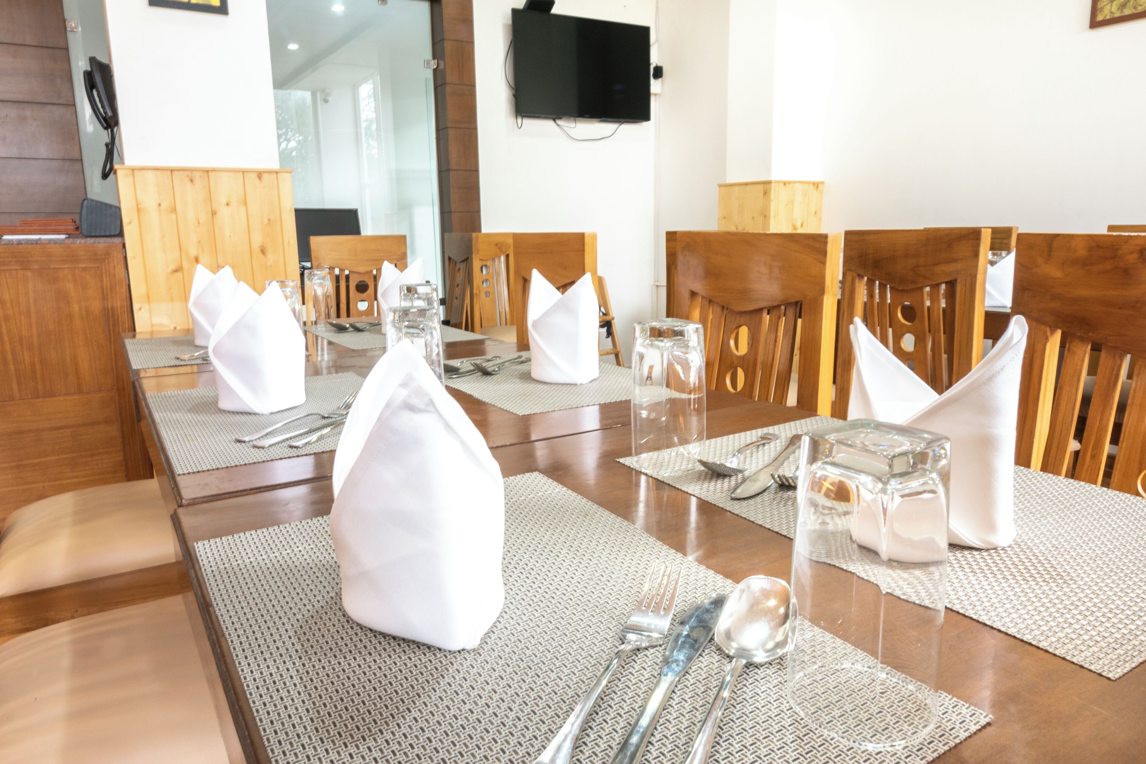 A dining table set with white napkins, cutlery, and glassware, with wooden chairs and a television mounted on the wall.