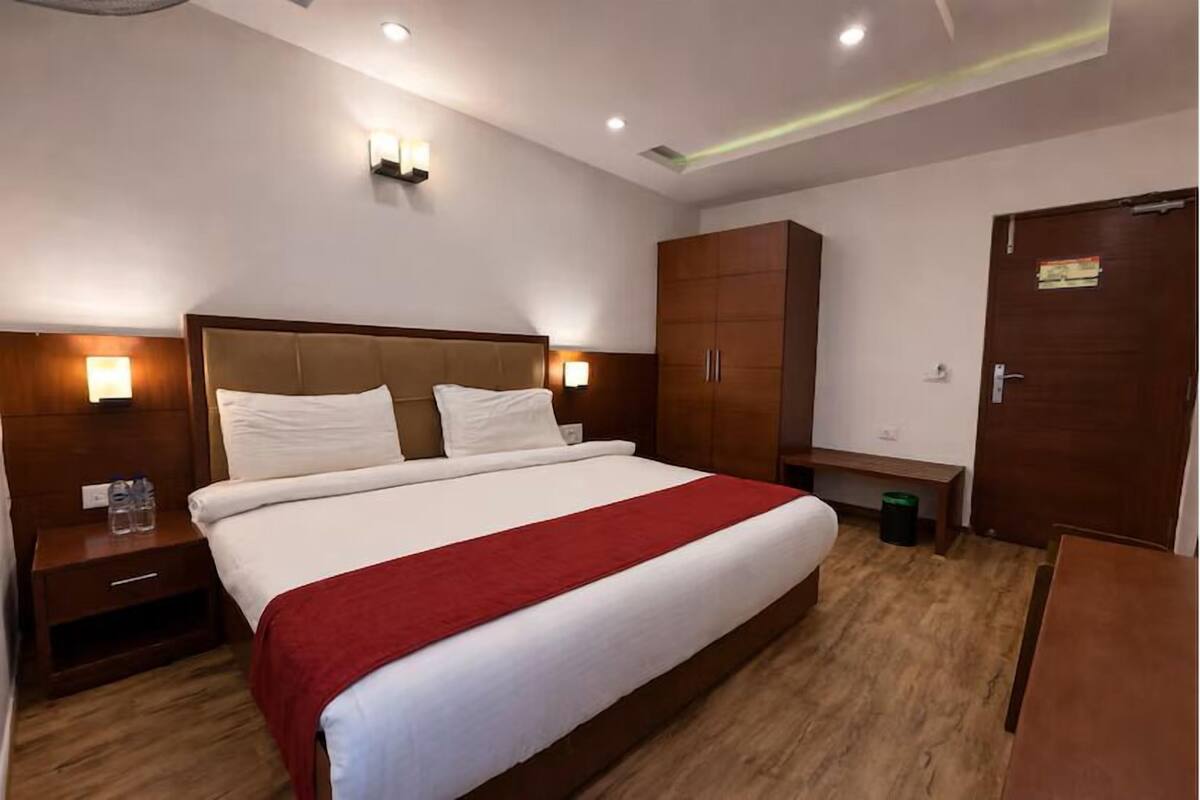 Habitación de hotel con una cama grande, cabecera de madera, dos mesitas de noche con lámparas, un armario y un pequeño banco.