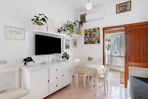 A room with a TV, white furniture, a dining table, chairs, and a bed.