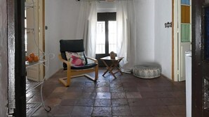 Une pièce comprenant une chaise, une petite table, un pouf rond, un luminaire suspendu en osier et une fenêtre avec des rideaux blancs.