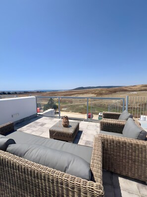 A rooftop terrace with wicker furniture, a small table, and a view of a coastal landscape.
