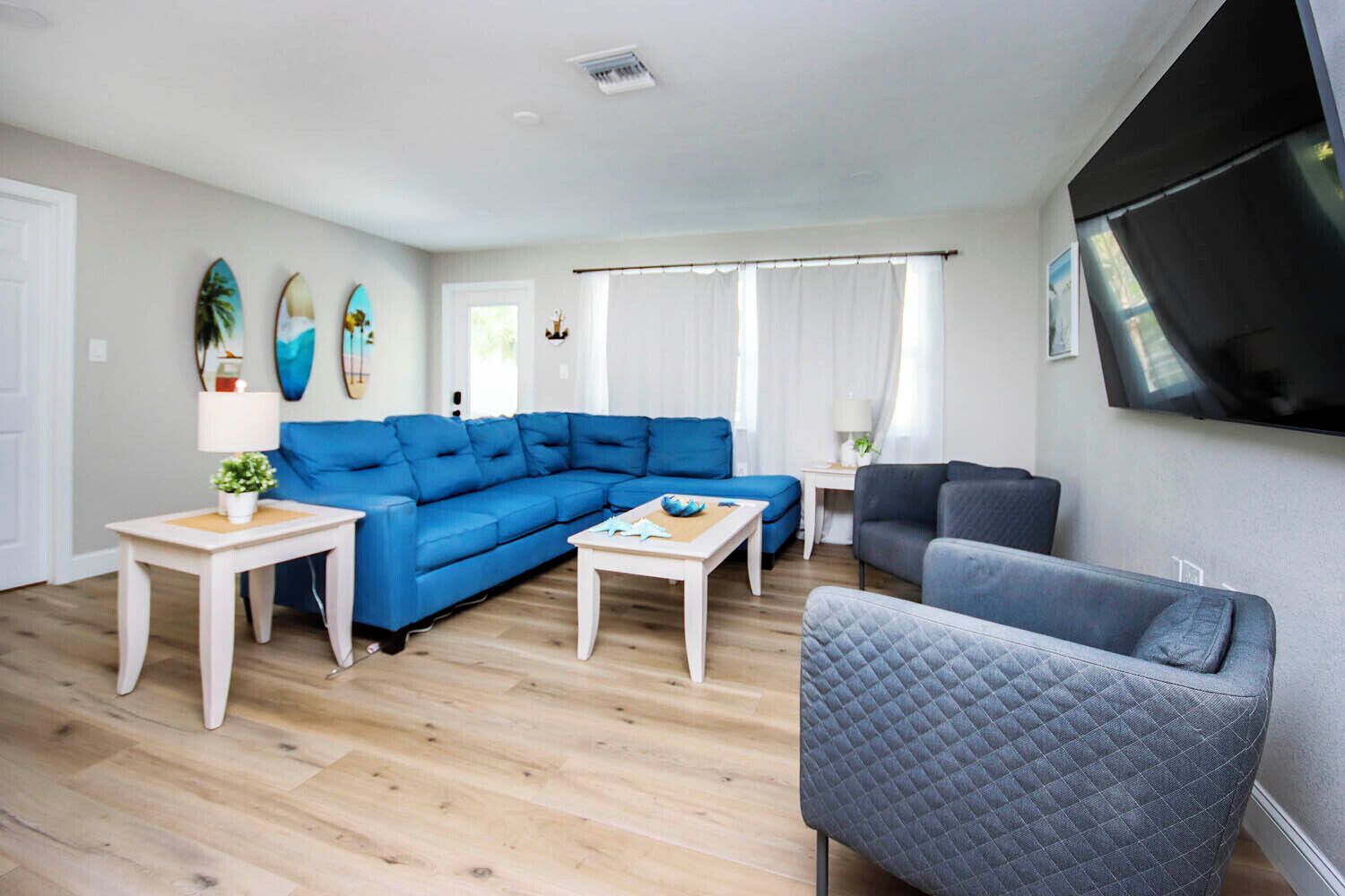 Un salon moderne avec un canapé d’angle bleu, un fauteuil gris et une petite table blanche. On peut voir des planches de surf accrochées au mur et un téléviseur à écran plat fixé au mur.