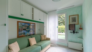 Una habitación con un sofá verde, un cuadro, un televisor y una ventana con vista a espacios verdes.