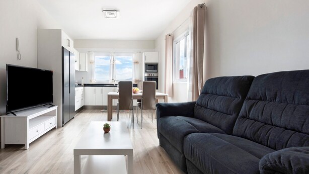 Una sala de estar moderna con un televisor de pantalla plana, un sofá gris oscuro, una mesa de centro blanca y una cocina al fondo.