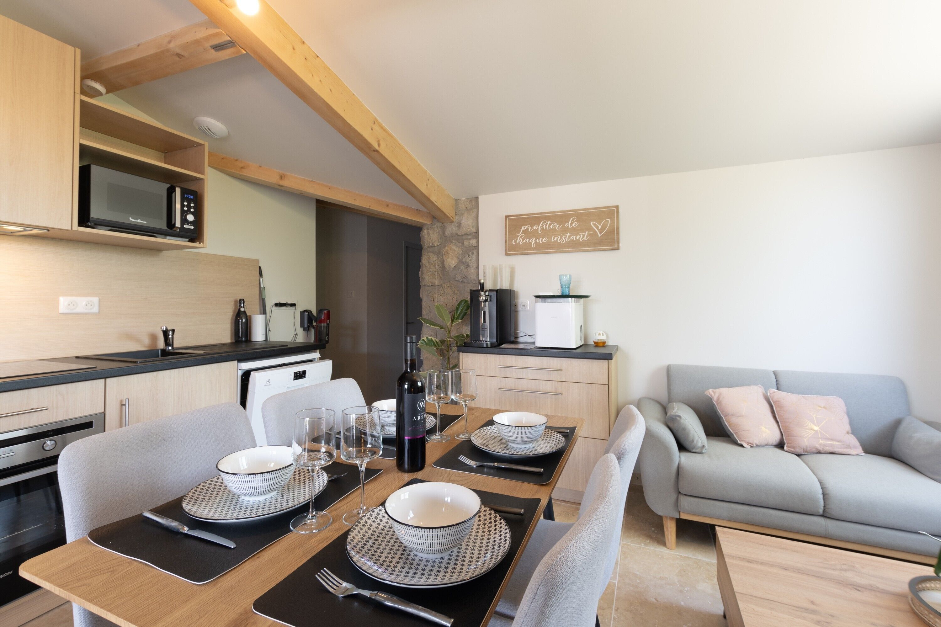 A modern kitchen with wooden beams, a dining area with a table set for two, and a grey sofa.