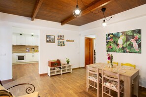 A spacious room with a wooden dining table, chairs, a kitchen in the background, and a ceiling with exposed beams.