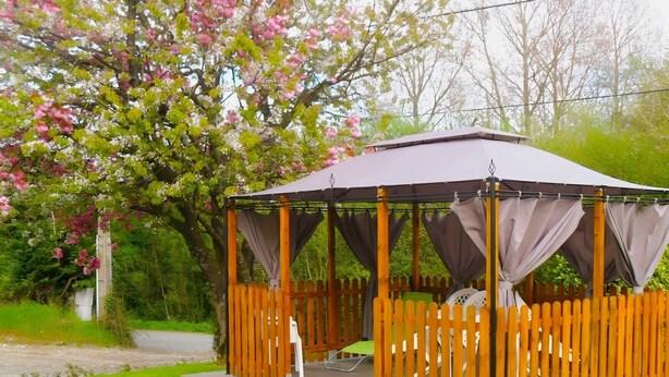 Ein hölzerner Pavillon mit Dach und Vorhängen, umgeben von einem Holzzaun, inmitten eines üppigen Gartens mit blühenden Bäumen.