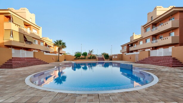 A swimming pool surrounded by a tiled deck, with multi-story buildings featuring balconies and steps leading to the pool.