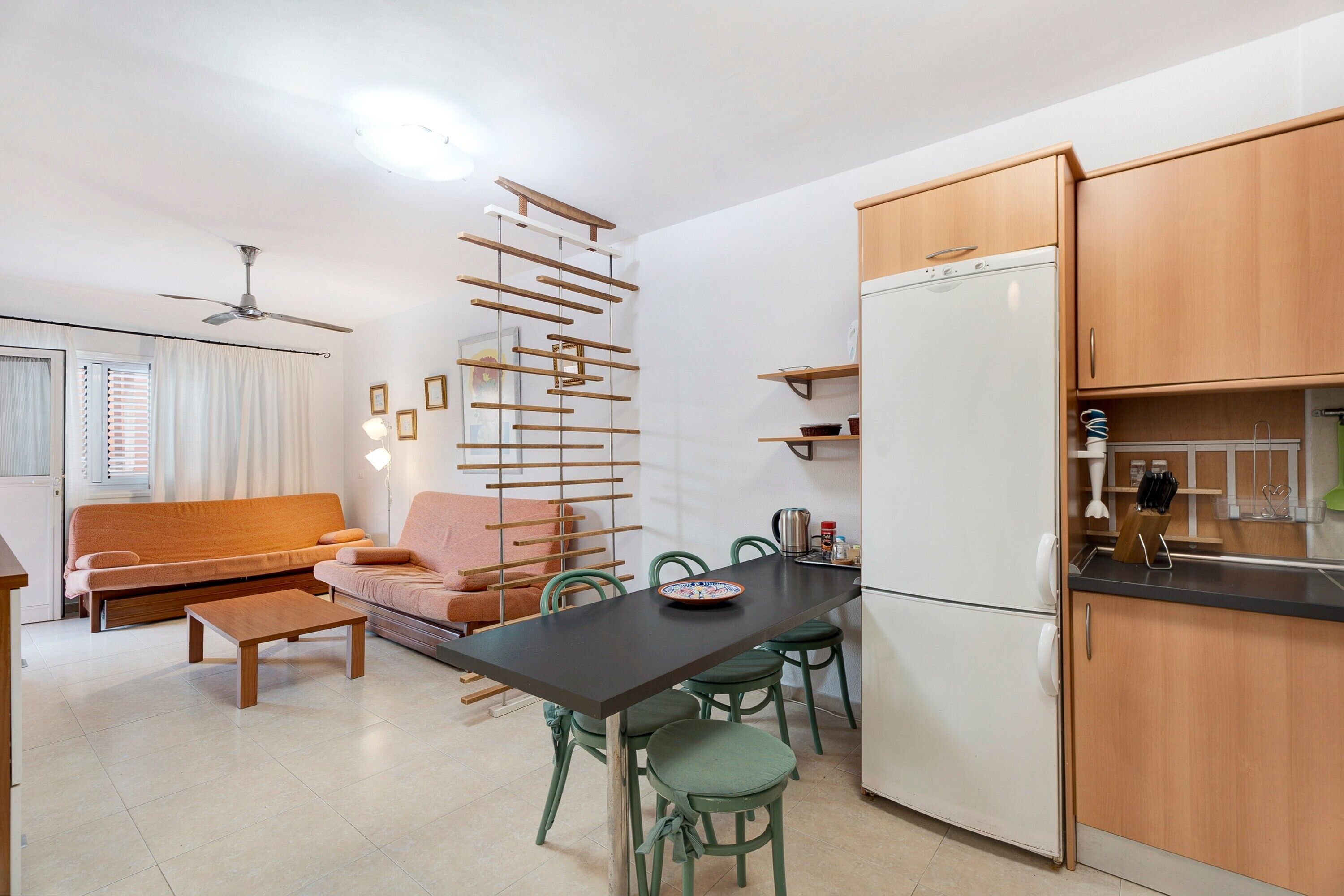Een compacte keuken met een koelkast, houten keukenkastjes, een eettafel met stoelen en een bank.