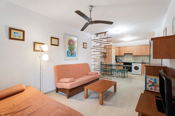 Un espace de vie compact comprenant un canapé, une table basse, un téléviseur, un ventilateur de plafond, un coin repas avec des chaises, une cuisine avec des placards et un lave-linge.