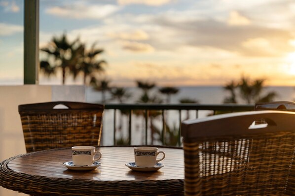 A wicker table with two cups and saucers, overlooking a body of water with palm trees.