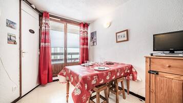 A dining area with a table set for two, a television on a wooden cabinet, and a balcony with red curtains.