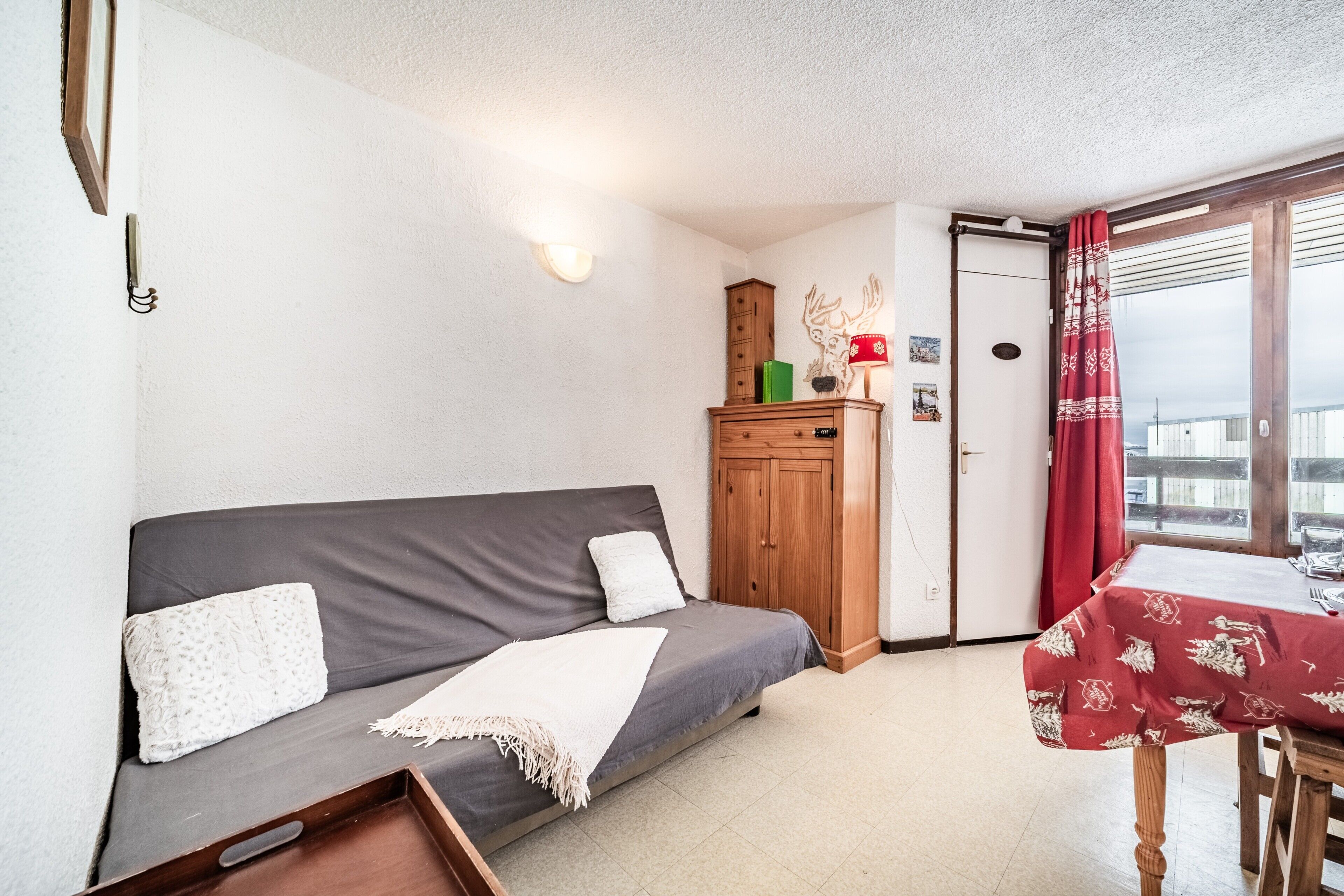 Une pièce avec un canapé gris, un meuble en bois, une table recouverte d’une nappe rouge et une fenêtre avec des rideaux rouges.