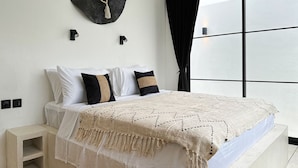 A modern bedroom with a large bed, white bedding, a decorative wall hanging, and a view through a large window.