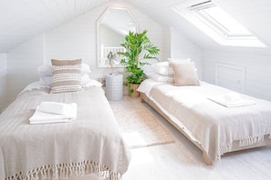 Two single beds with white linens and matching pillows, a potted plant, and a side table create a peaceful retreat.