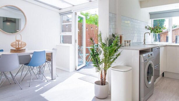 A modern kitchen with a built-in washing machine, a dining area with a table and chairs, and a large window providing natural light. - Stunning Townhouse with Large Garden - Sleeps 8 (Cheltenham)