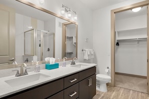 A modern bathroom with a large mirror, two sinks, a toilet, and a walk-in closet.