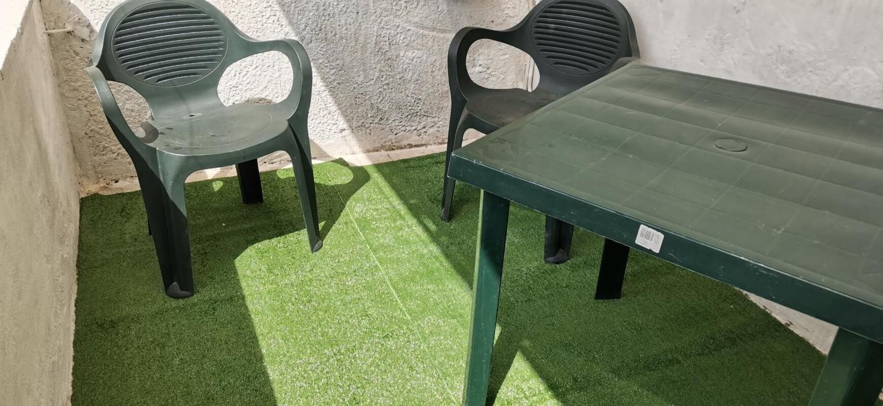 En liten sittplats utomhus med ett grönt plastbord och två stolar, omgiven av en betongmur och konstgjort gräs.