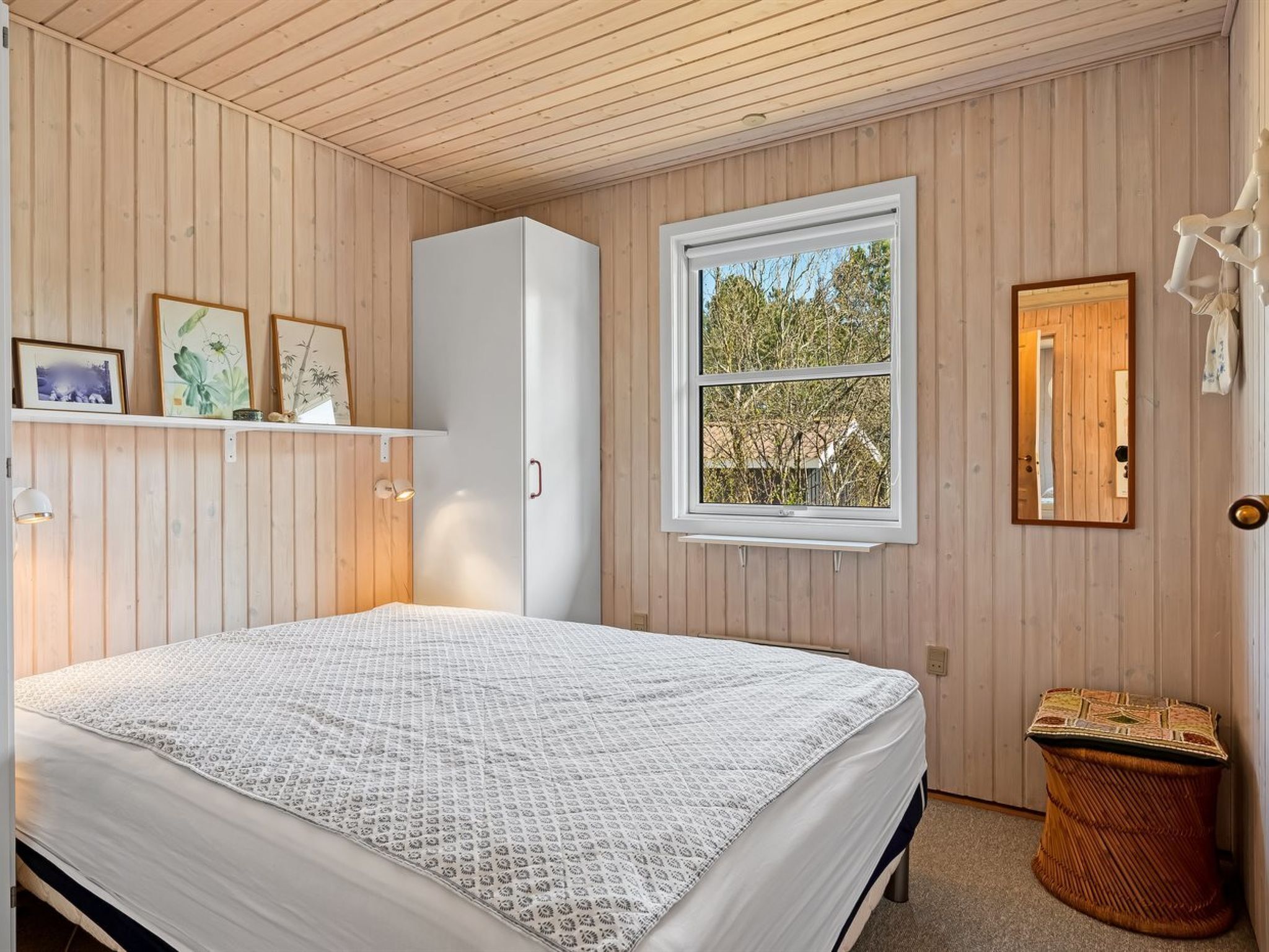 A bedroom with a bed, wooden walls, a window with a view, a wardrobe, and a small stool.