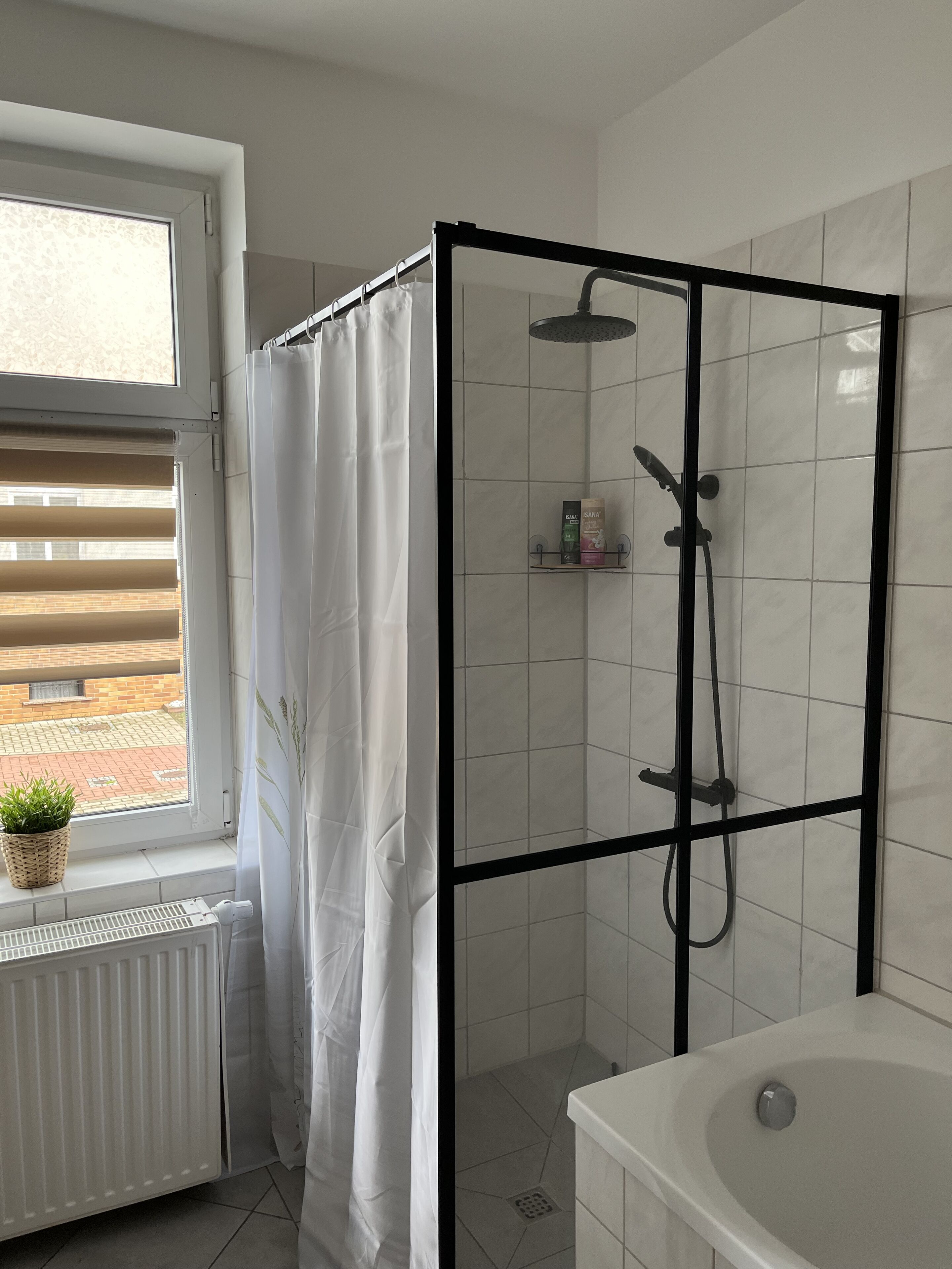 A modern bathroom with a glass-enclosed shower, a white bathtub, and a window with blinds.