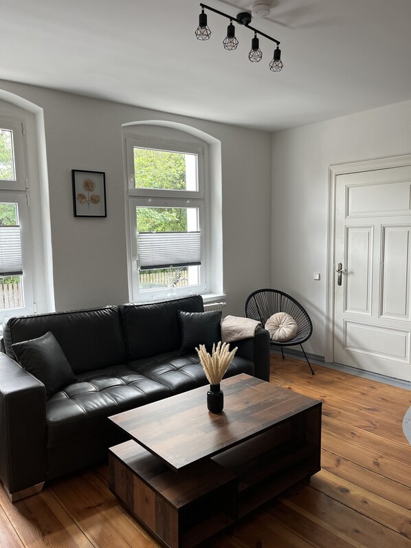 Un salon moderne avec un canapé en cuir noir, une table basse en bois et un fauteuil noir. Il y a deux fenêtres équipées de stores, une porte blanche et un luminaire au plafond.