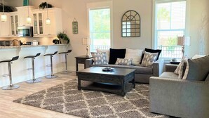 A modern living room with a grey sofa, a coffee table, and a patterned rug. There is a kitchen area with white cabinets and a microwave. - 🦩Book Me Baby! 30A Oasis~Private Heated Pool~Pet-Friendly🦩 (Santa Rosa Beach)