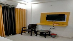 Una habitación con cama, una silla, una mesita, un televisor en un estante amarillo y una cortina.