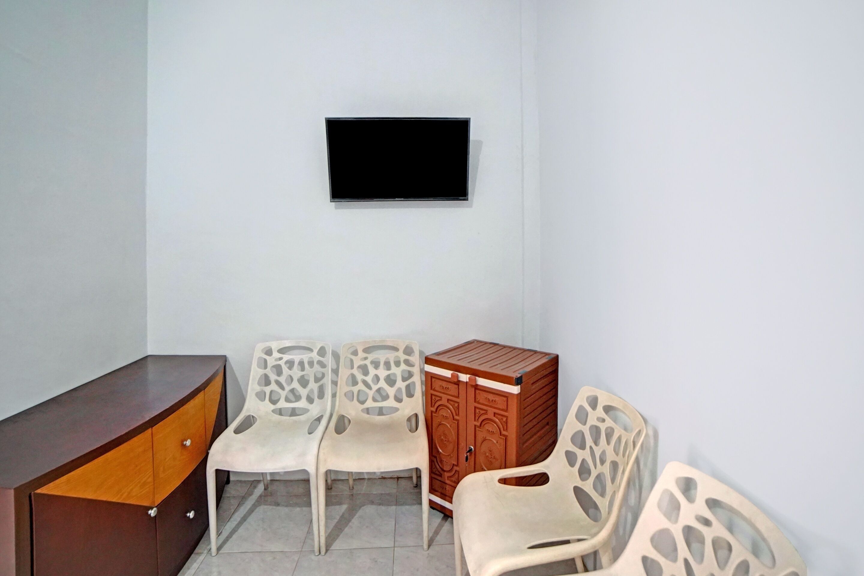 Ein Zimmer mit einem an der Wand befestigten Flachbildfernseher, einem Holzschrank und drei weißen Plastikstühlen.