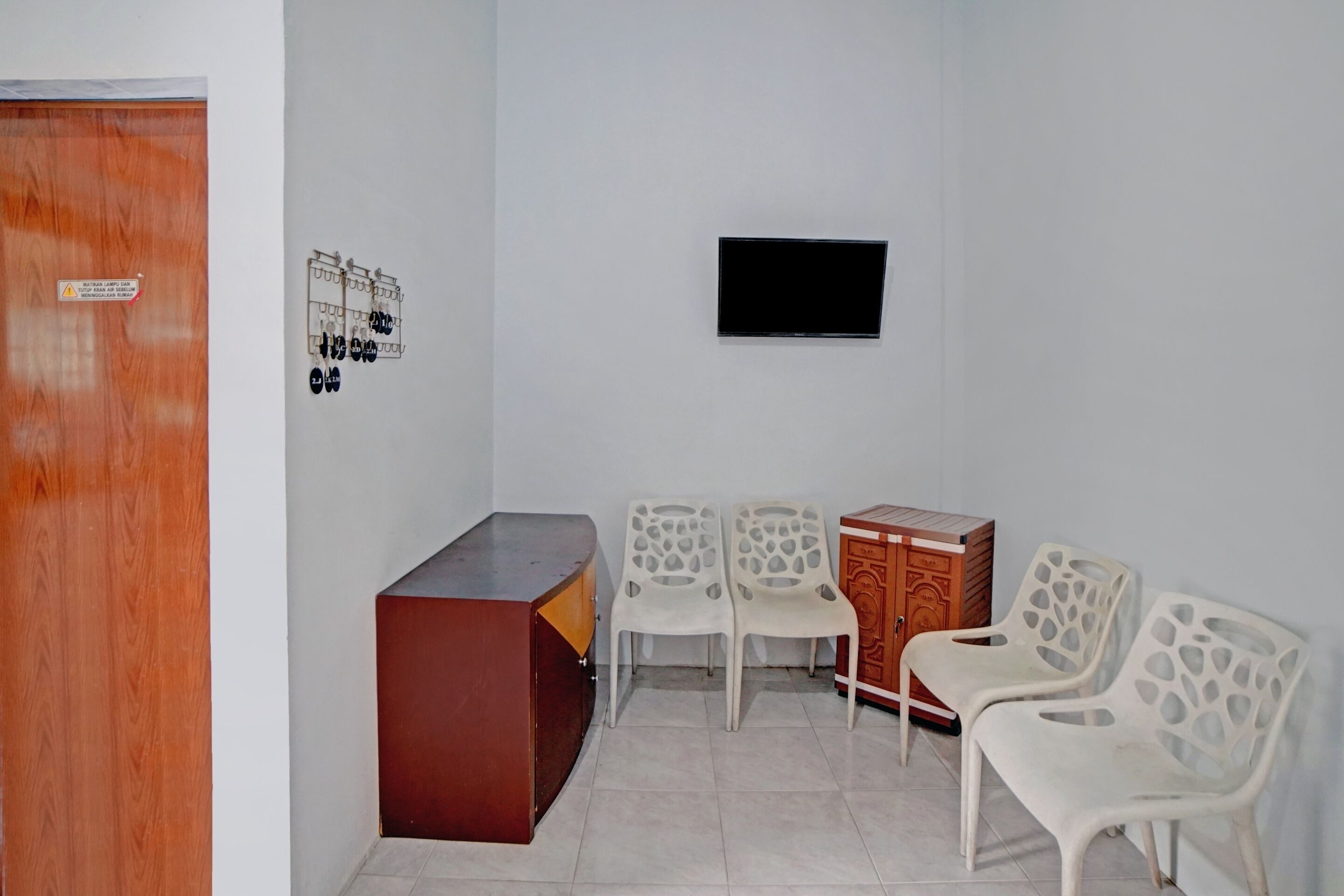 A room with a wooden door, a TV mounted on the wall, a small wooden cabinet, and four white plastic chairs.