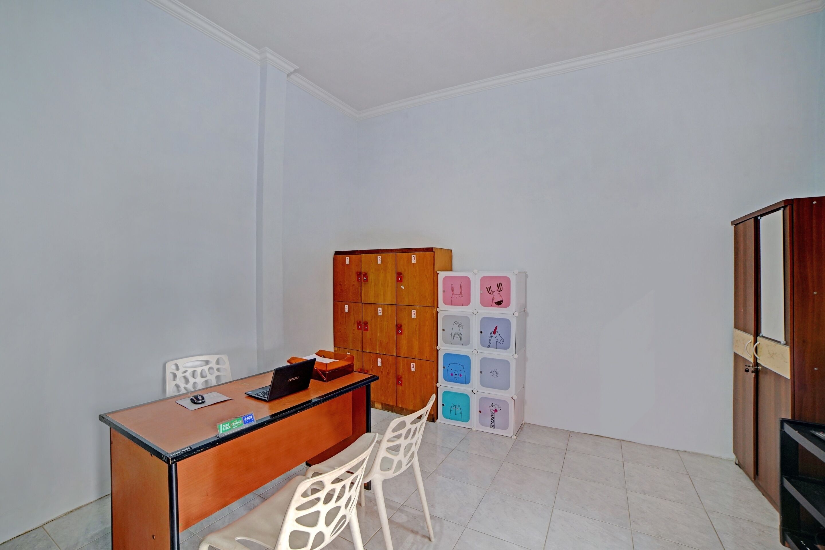 Une petite pièce avec un bureau en bois, deux chaises blanches, un meuble avec plusieurs compartiments et un tableau blanc orné de dessins.