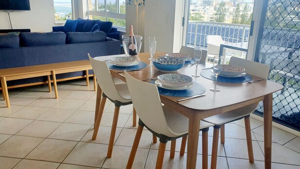Zona pranzo con tavolo imbandito per quattro, sedie, una bottiglia di champagne e vista sulla città attraverso ampie finestre.
