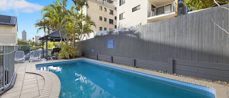 A swimming pool with a wooden deck, lounge chairs, and a fenced area.