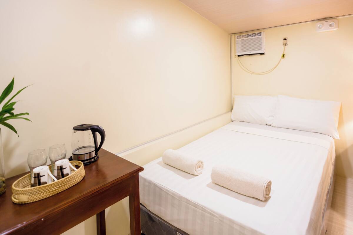 A hotel room with a bed, a bedside table with a kettle, glasses, and a plant.