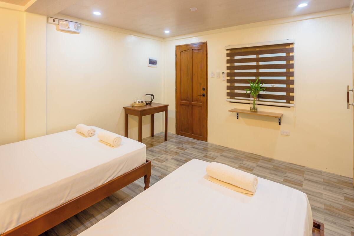 A hotel room with two single beds, a wooden nightstand, a small table with a kettle, a window with blinds, and a wall-mounted shelf with a plant.