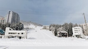 A snowy urban street with buildings, including a multi-story hotel and a two-story residential building. - Hotel Justice (Minamiuonuma)
