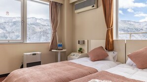 A hotel room with two beds, a bedside table with a lamp, a window with curtains, an air conditioning unit, and a view of snow-covered mountains. - Hotel Justice (Minamiuonuma)