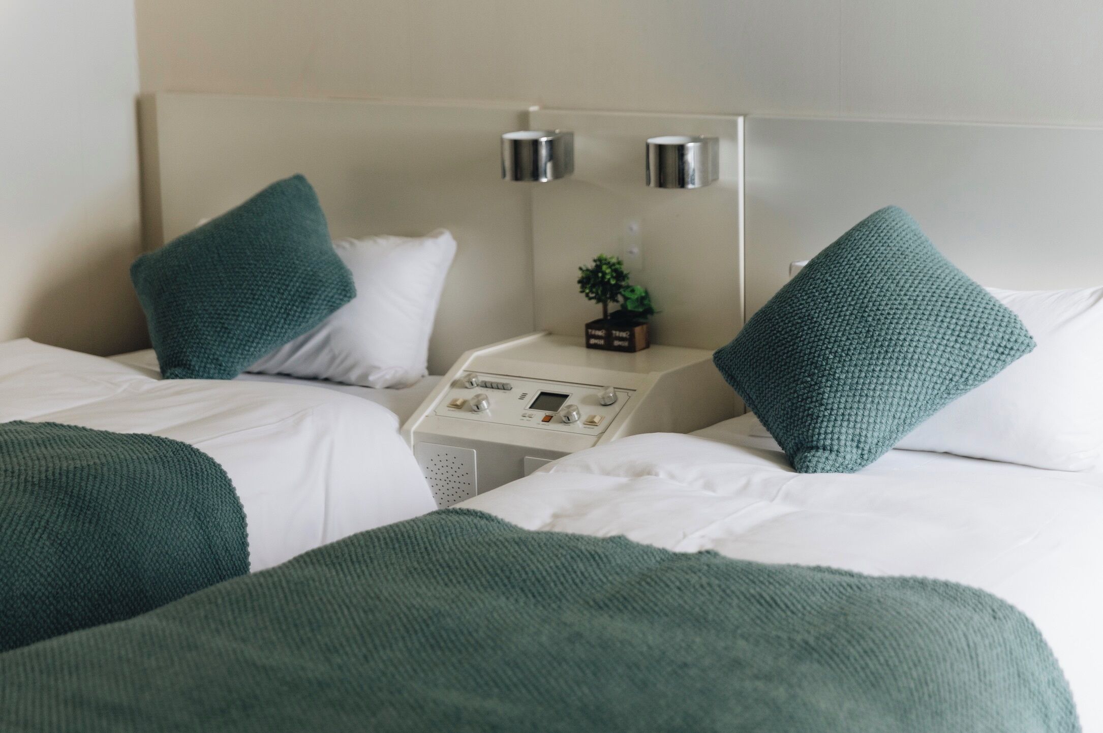 A hotel room with two beds, each with a green knitted throw, white bedding, and a bedside table with a small plant and a control panel.