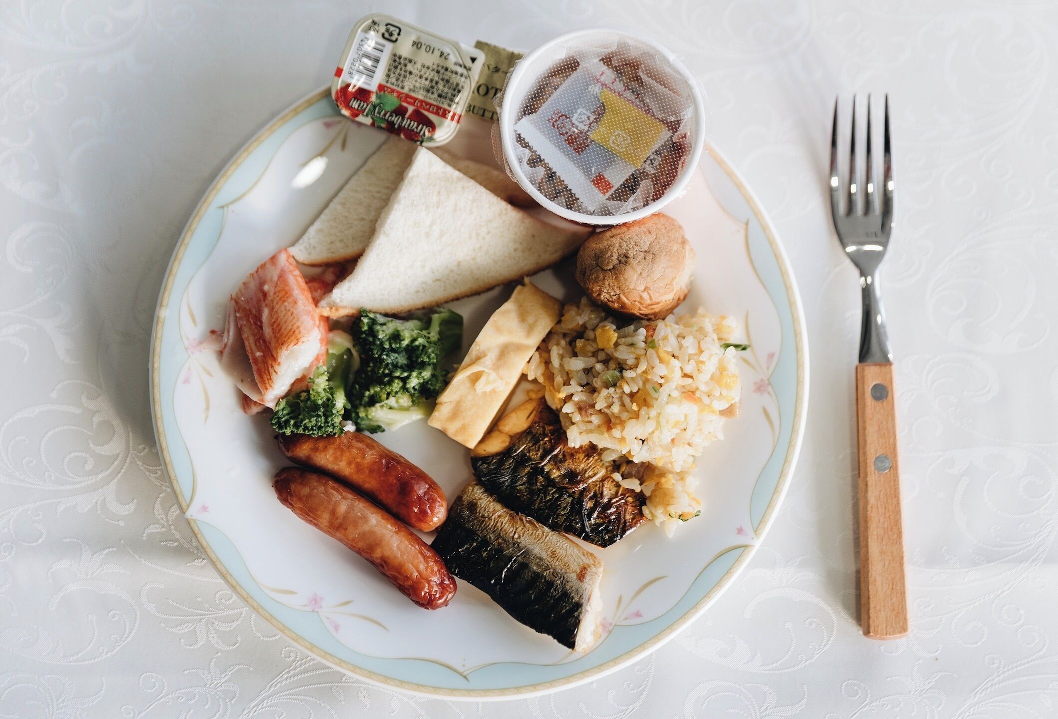 Un plato con diversos alimentos, como salchichas, pan y arroz, acompañado de una taza de té y un tenedor sobre una mesa con mantel estampado.