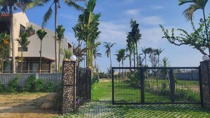 Una entrada con reja y un camino pavimentado que lleva a un edificio de diseño moderno, rodeado de palmeras y un cielo despejado.