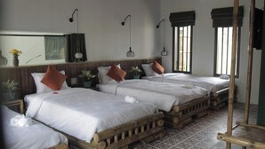 Habitación de hotel con tres camas de madera, ropa de cama blanca y almohadas marrones. Hay lámparas fijadas a la pared y un ventanal con cortinas.