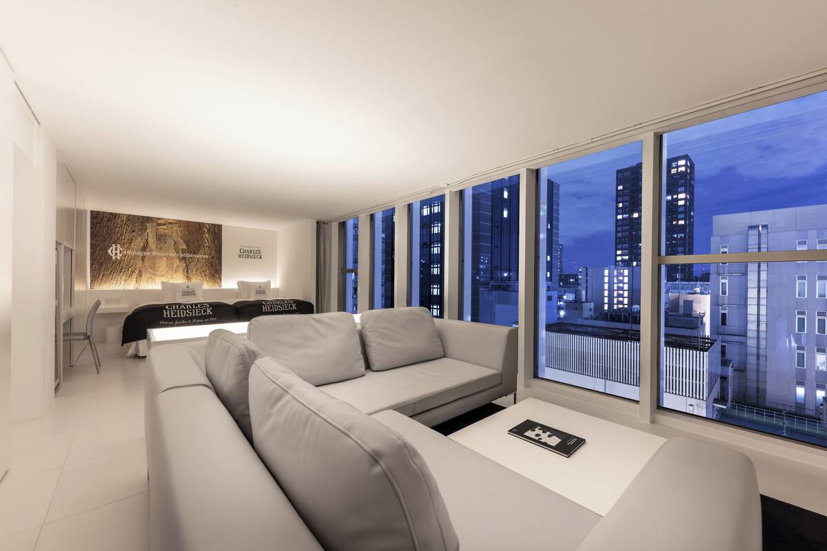 A modern living room with a sofa, a coffee table, and a dining area with a table and chairs. Large windows offer a view of the cityscape at night.