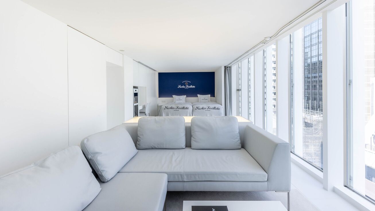 Une chambre d’hôtel moderne avec un canapé gris, une petite table et une grande fenêtre donnant sur la ville.