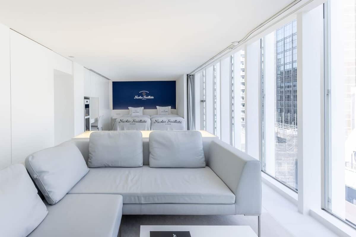 A modern hotel room with a grey sofa, a small table, and a large window offering a city view.
