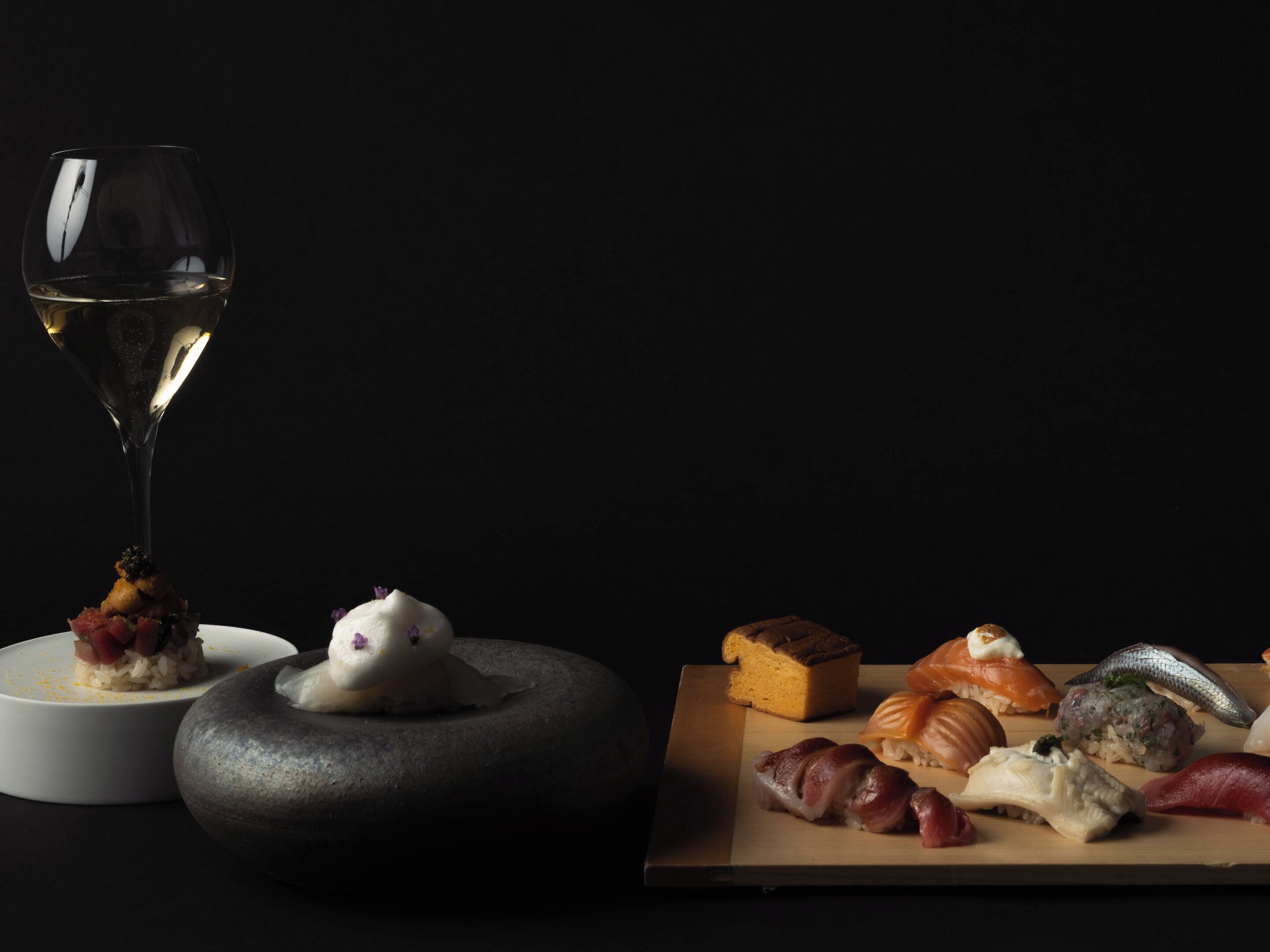 Une variété de sushis présentés sur une planche en bois, un verre de vin blanc et un bol en pierre.