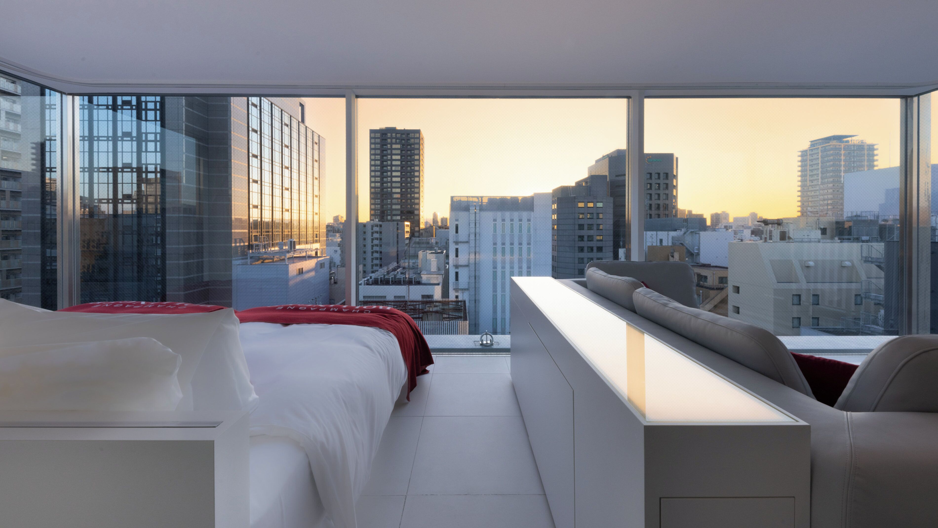 A modern hotel room with a large window offering a city view, a bed, a sofa, and a small table.