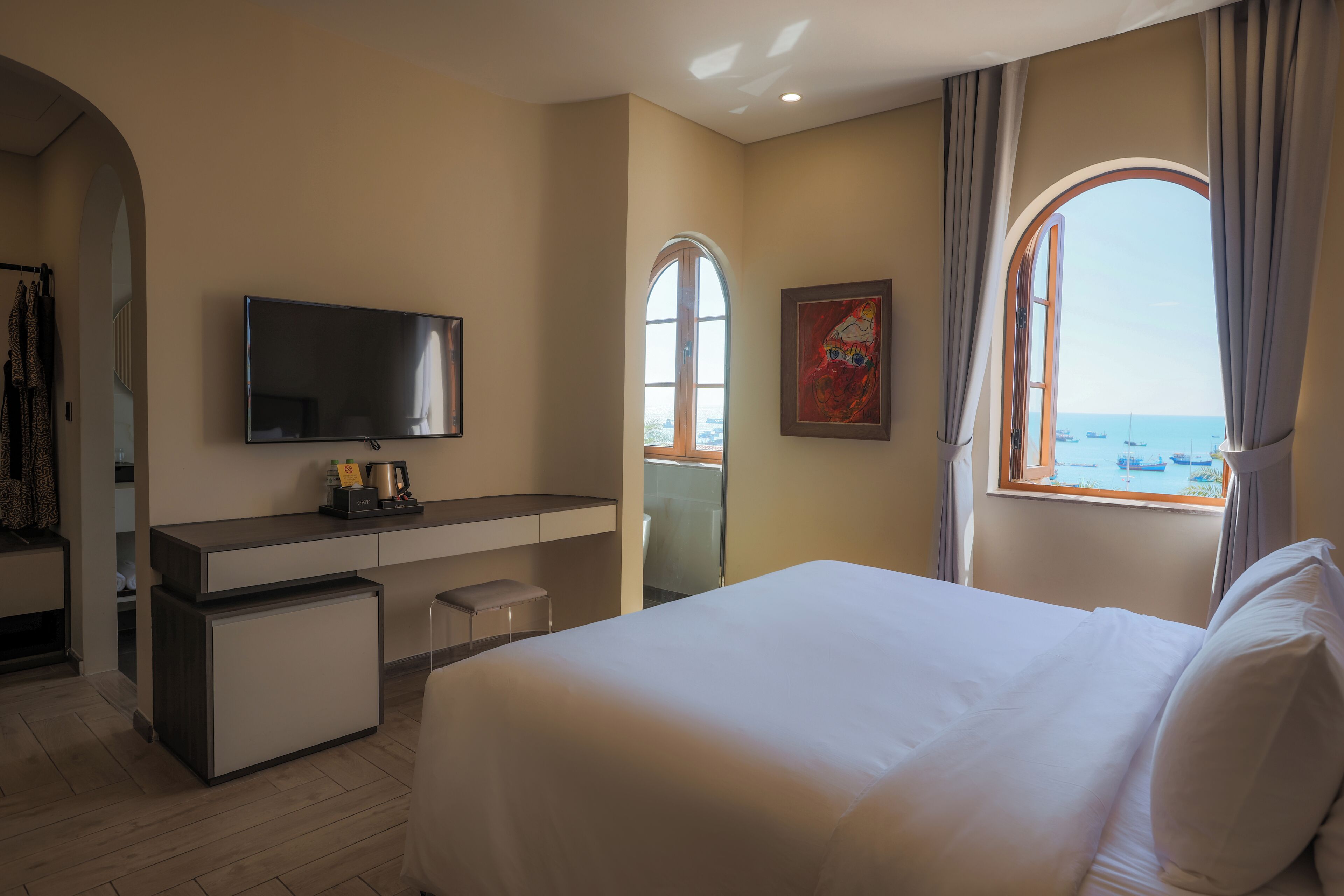 Een hotelkamer met een bed, een bureau, een televisie, een kleine koelkast en een raam met uitzicht op zee.