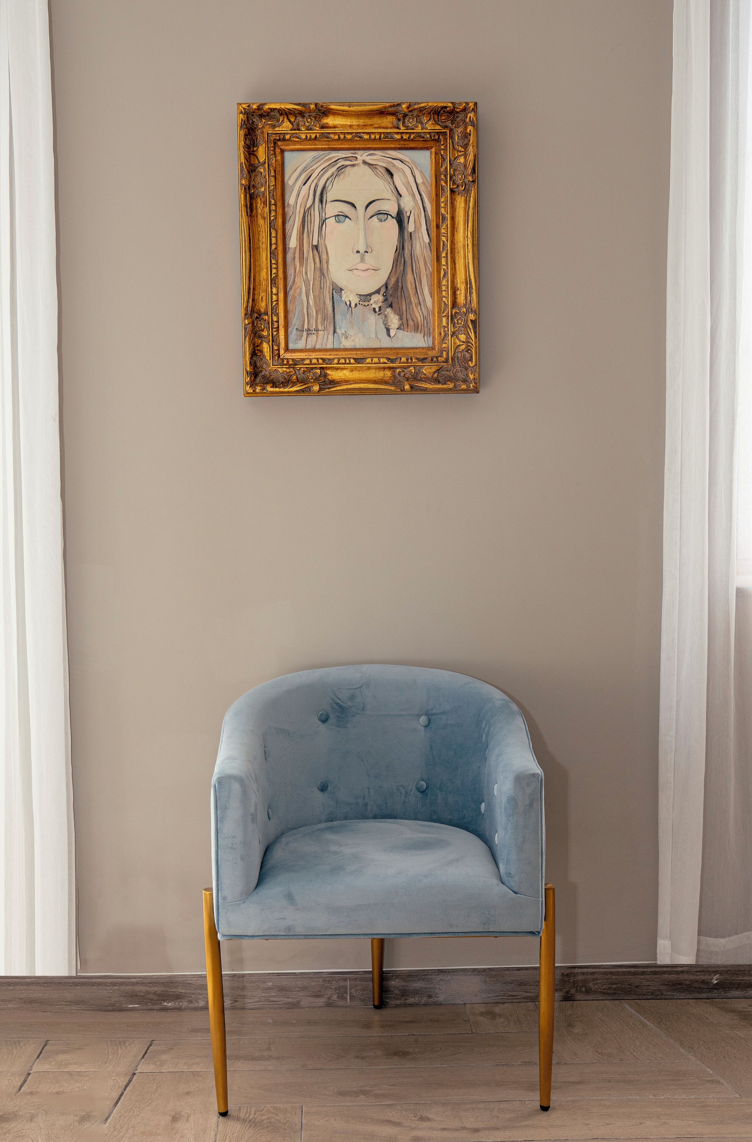 A light blue armchair with wooden legs, positioned against a wall with a framed artwork above it.