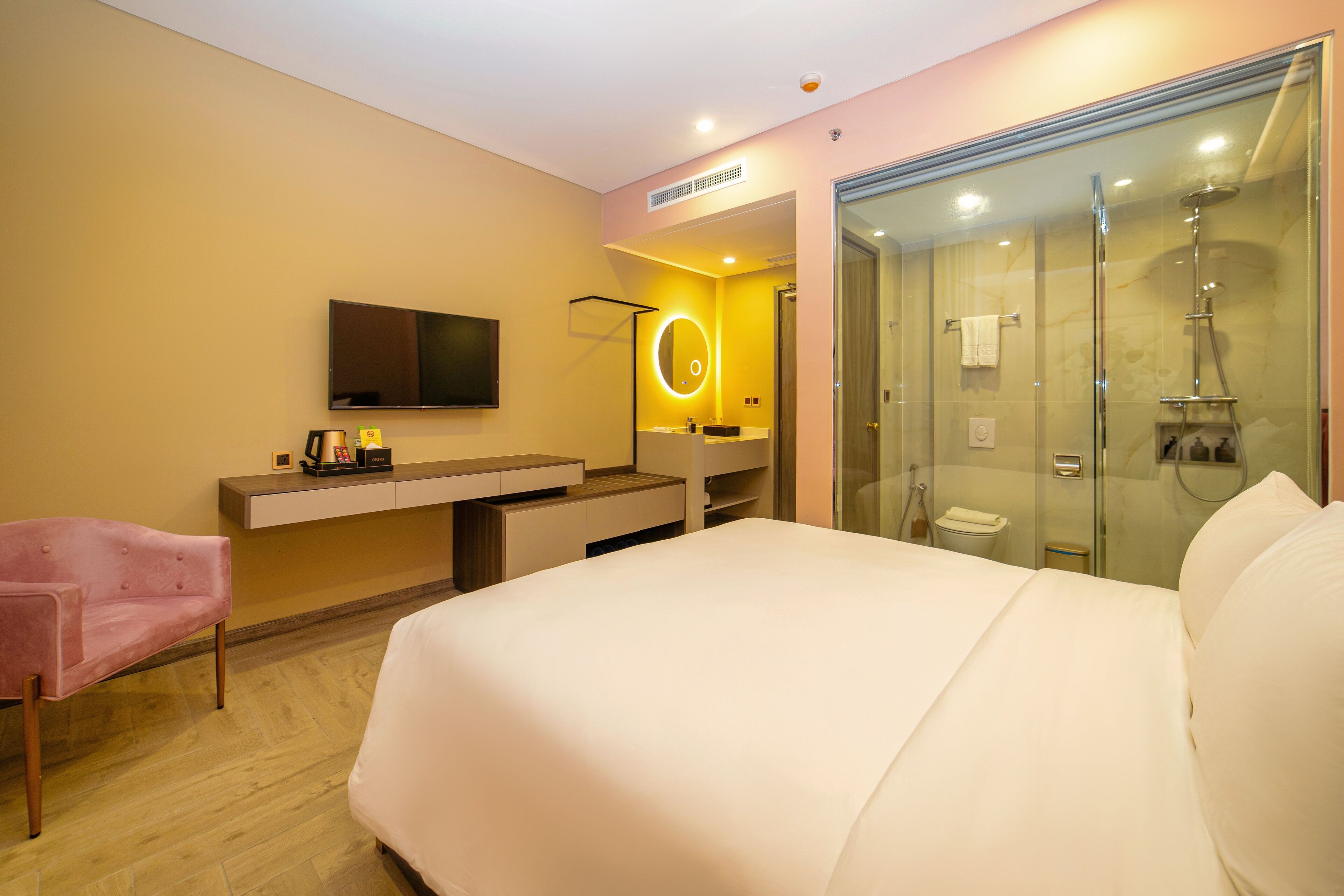 Une chambre d’hôtel moderne dotée d’un grand lit, d’une coiffeuse avec miroir, d’une télévision et d’une douche séparée par des parois en verre.