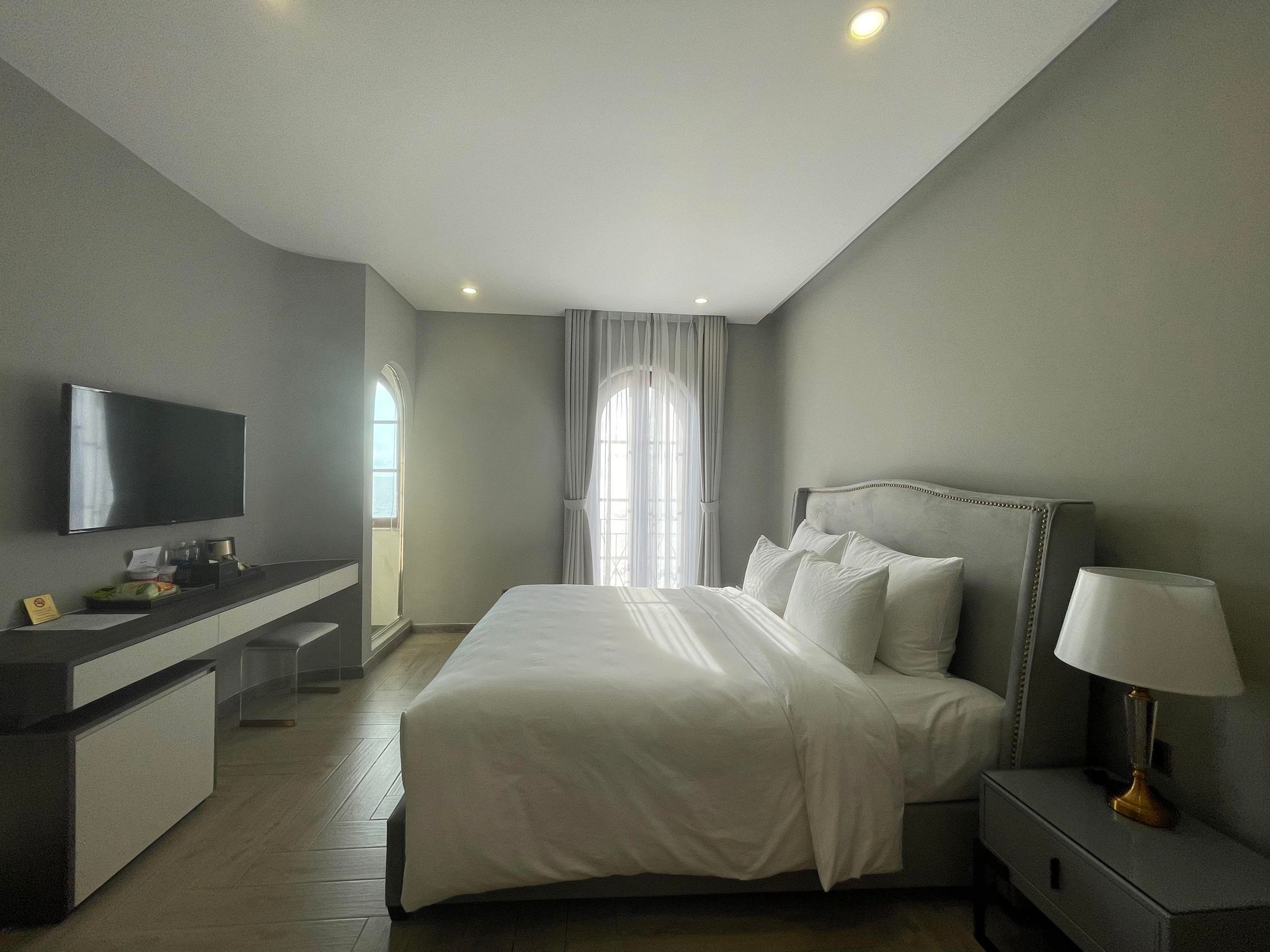 A modern hotel room with a large bed, a bedside table with a lamp, a TV mounted on the wall, and a desk with a chair.
